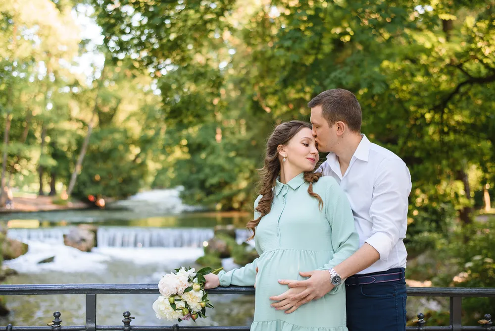Couple in casual attire shares a tender moment by a river with a small waterfall, surrounded by lush greenery. Woman holds a bouquet of white flowers.