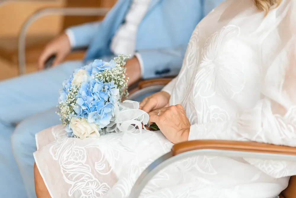 Bride in white lace gown holds blue hydrangea bouquet with baby's breath, seated beside groom in light blue suit. Hands clasped together.