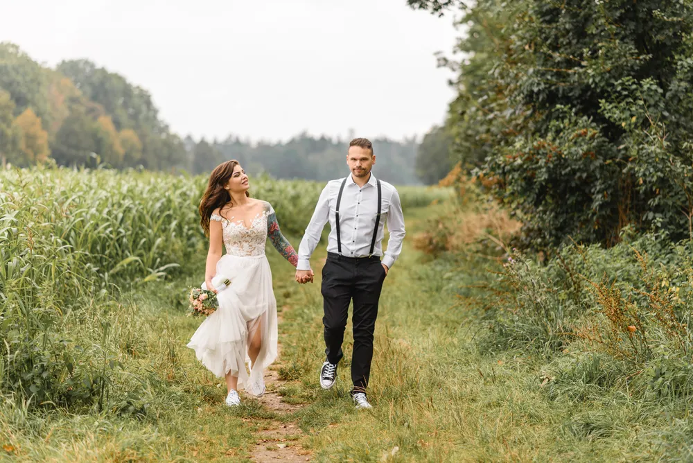 Couple in wedding attire walks hand-in-hand along a rural dirt path surrounded by tall grasses and trees on a cloudy day.
