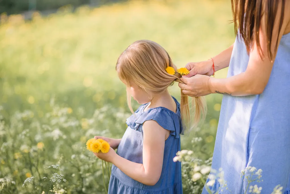 A woman adorns a young girl's hair with dandelions in a sunlit meadow, both wearing blue dresses amidst a field of wildflowers.