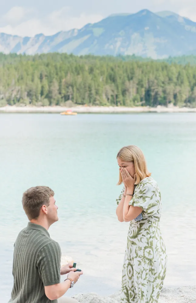 A man kneels by a lake, offering an engagement ring to a tearful woman in a floral dress, with mountains and a boat in the background.