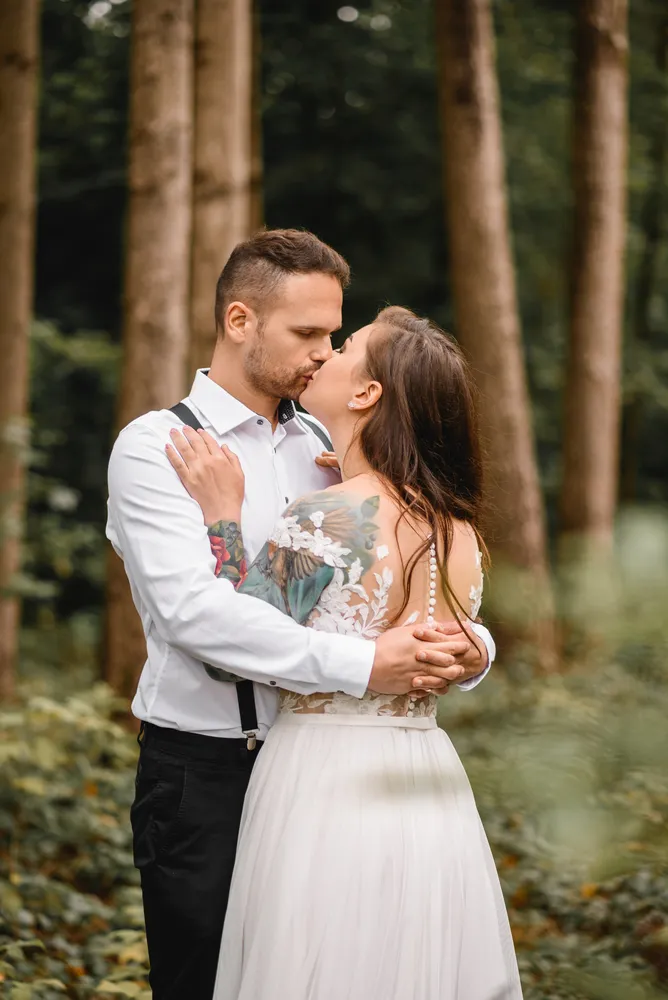 Couple in forest, sharing kiss. Man in white shirt and black pants, woman in white lace dress. Tattoos visible on woman's arm. Tall trees and greenery in background.