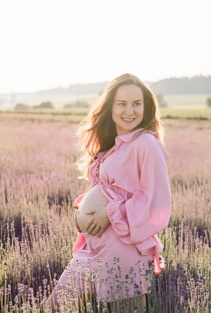Expectant mother in pink dress stands in lavender field, cradling belly, smiling softly, golden hour lighting.