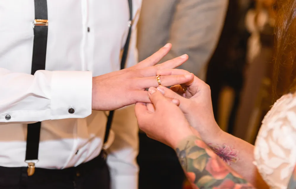 Individual in white shirt with black suspenders receiving a ring from another person with floral tattoos, during a formal event.