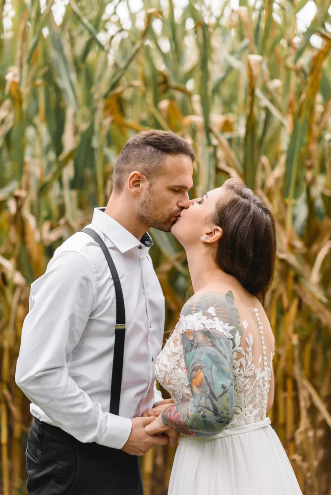 Couple in wedding attire kissing outdoors with corn stalks in the background.
