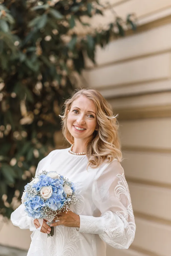 Woman in white lace dress holds blue and white bouquet, standing outdoors with blurred foliage and building in background.
