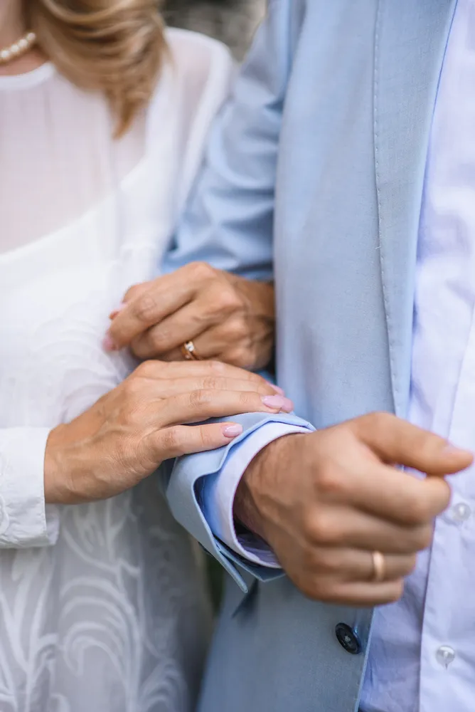 A man and woman, dressed in formal attire, stand closely together. Their hands are gently clasped, suggesting a bond or partnership. The woman's white dress with lace details and the man's light blue suit with a matching shirt are in focus, conveying a sense of elegance and formality. The background is indistinct, keeping the attention on the subjects and their connection.