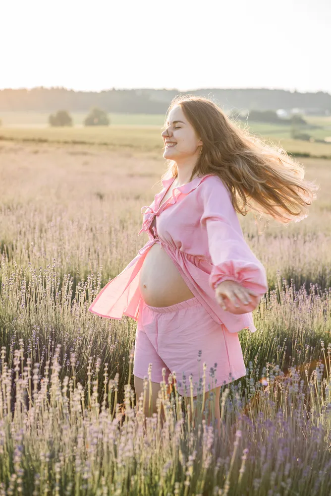 Expectant woman in pink jumpsuit joyfully twirls in lavender field at golden hour.