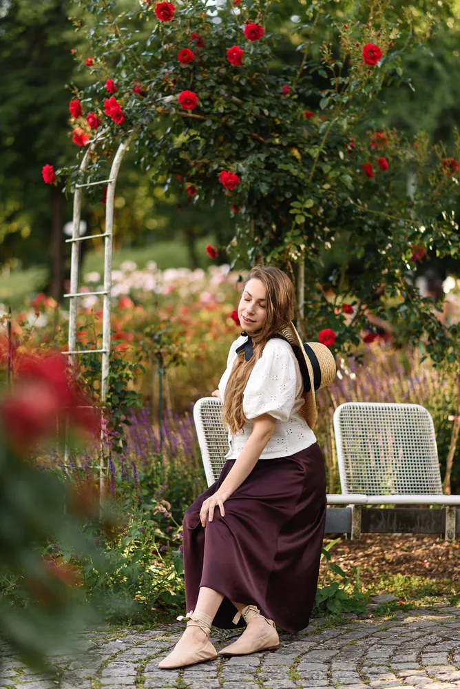 Woman in white blouse and maroon skirt sits on cobblestone path, surrounded by red roses and greenery in a sunlit garden.