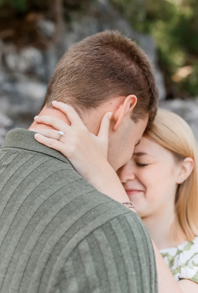 Man and woman embracing outdoors. She wears a ring, he has short hair. Both smiling. Blurred natural background.
