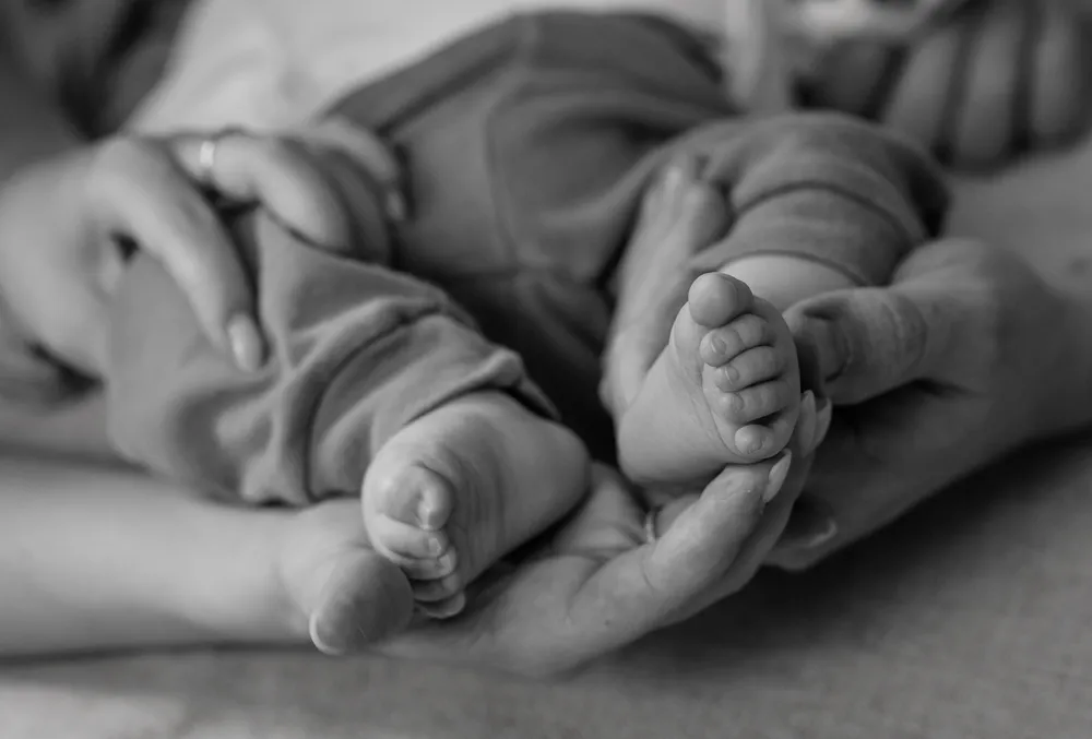 Two hands gently cradle a baby's feet in a monochrome close-up, highlighting the soft skin and tiny toes of the infant. The adult's fingers, adorned with a ring, carefully support the child's ankles and feet, creating an intimate and tender moment.
