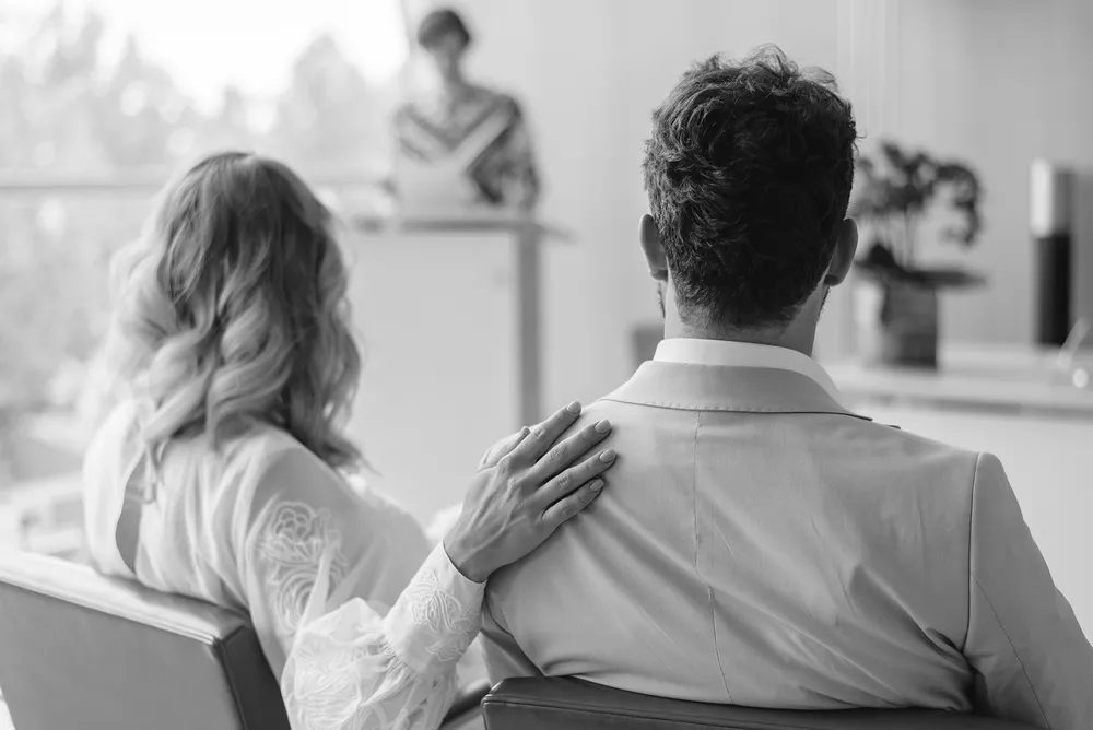 Man and woman seated at conference, her hand on his shoulder, presenter speaking at podium, modern office setting.