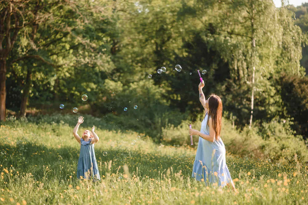 Two young girls in a sunny meadow play with soap bubbles, one reaching up, the other blowing them with a wand, both wearing blue dresses.