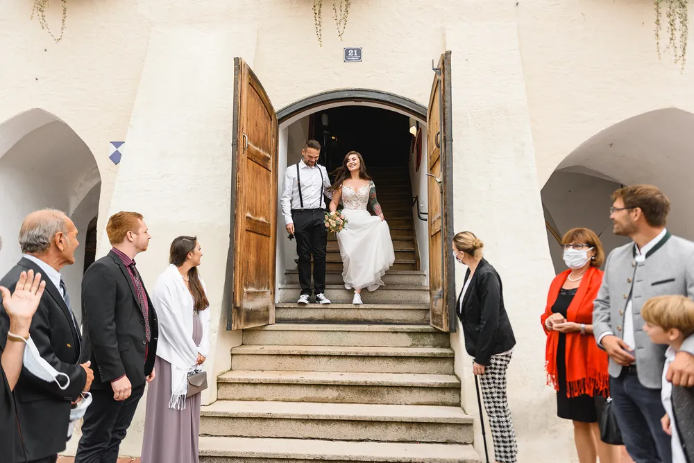 Bride and groom in modern attire descend stone steps flanked by guests, some wearing masks, outside a white-walled building with arched doorways, during a daytime wedding ceremony.