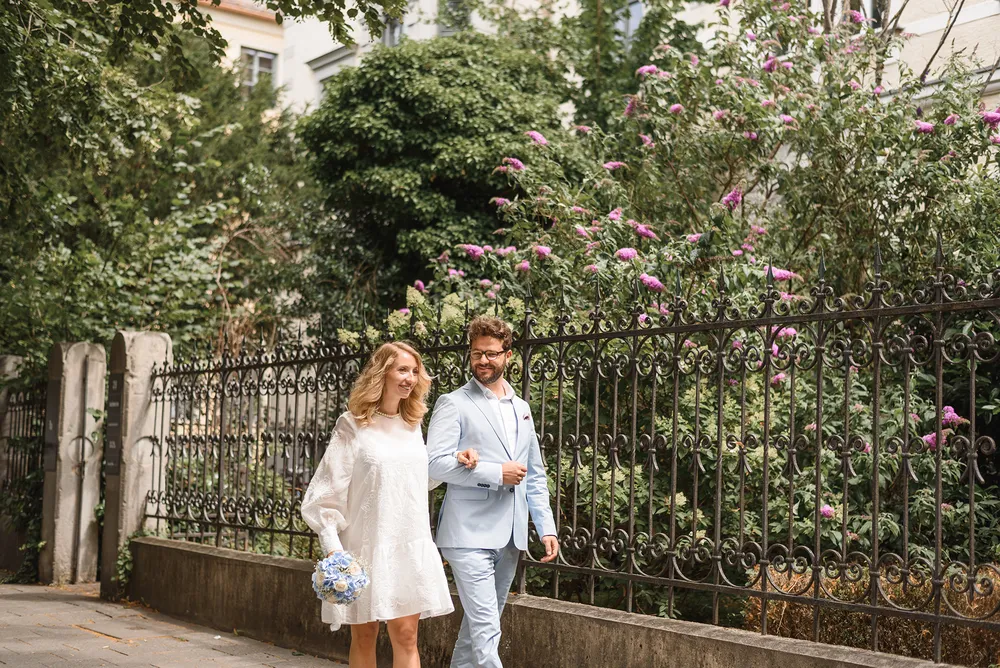 A couple in wedding attire walks hand-in-hand on a cobblestone path, surrounded by lush greenery and pink flowers, with an ornate black wrought-iron fence lining their route.