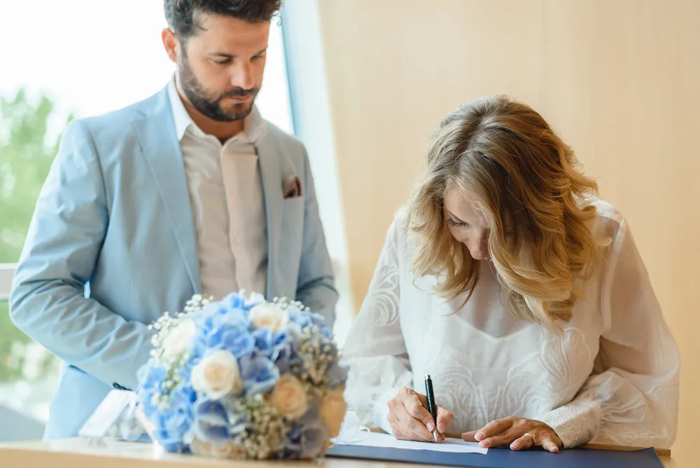 A man and woman in formal attire sign documents at a table, with a bouquet of blue and white flowers between them, indoors with natural light.