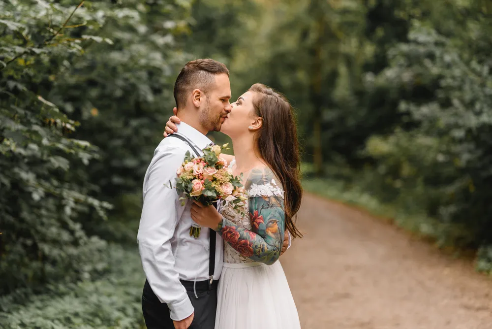 Couple in wedding attire shares a kiss on a forest path; bride holds bouquet, groom in suspenders.