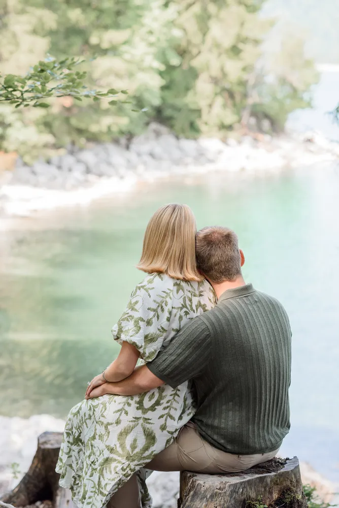 Couple embracing on tree stump by serene lake, surrounded by lush greenery. Soft focus background enhances intimate moment.