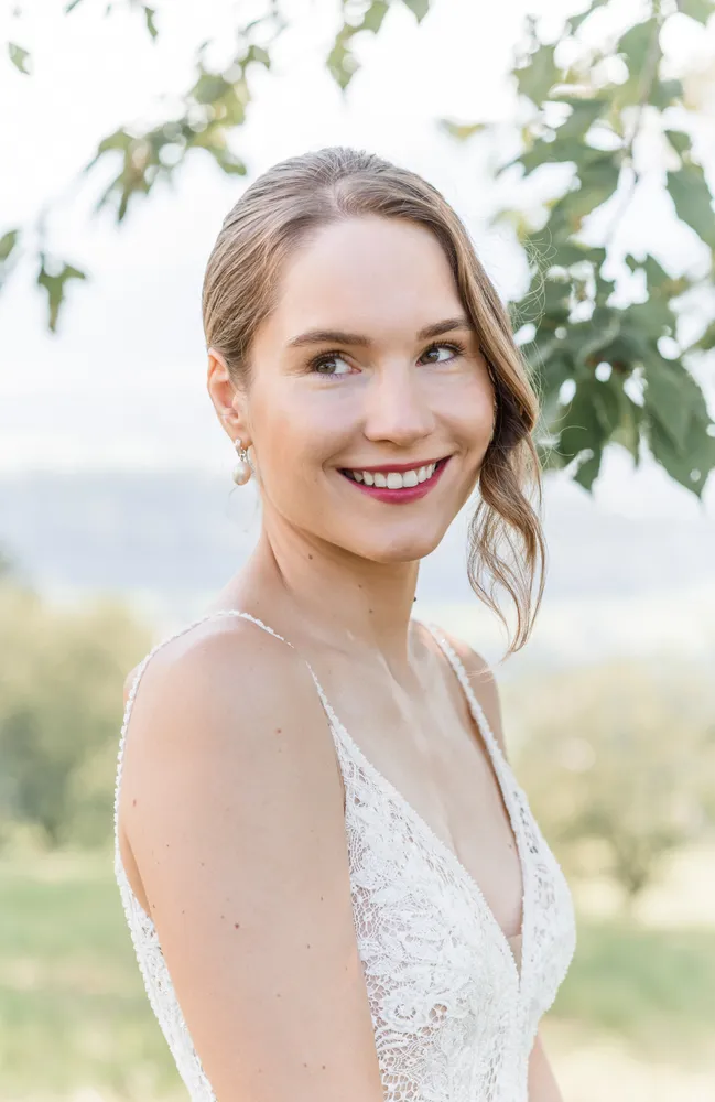 Woman in lace wedding dress smiles outdoors, with natural backdrop and soft lighting.