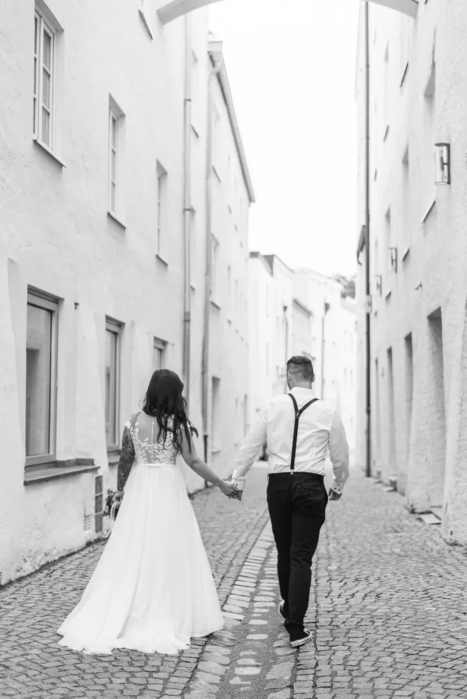 A couple strolls hand-in-hand down a cobblestone alley, bride in a flowing gown, groom in a white shirt with suspenders. Black and white photo, buildings on either side.