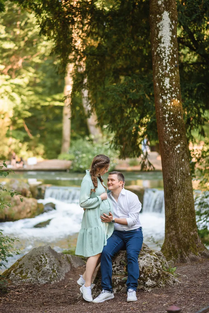 Pregnant woman in light green dress and man in white shirt and blue jeans embrace by a serene stream, surrounded by lush forest and soft sunlight filtering through trees.