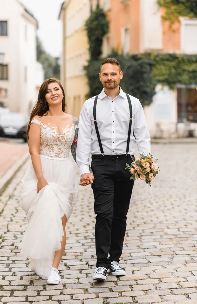 Couple strolls hand-in-hand on cobblestone street. Woman in white lace dress with floral bodice, man in white shirt and suspenders holds bouquet. Urban backdrop, daytime.