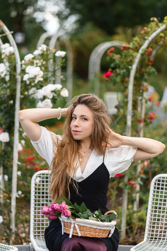 Woman with long brown hair sitting in a garden, holding a basket of pink roses, wearing a black and white dress, white metal chairs and trellises with white and red flowers in the background.