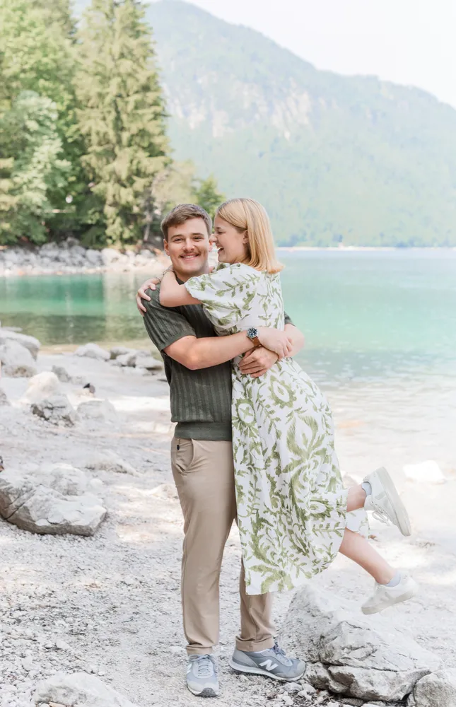 A couple joyfully embracing by a tranquil lake, surrounded by rocky terrain and lush greenery. The man wears a dark sweater and khaki pants, while the woman is in a floral dress. Both are smiling, exuding happiness in this natural setting.