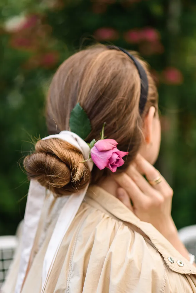 Woman's bun adorned with pink rose and green leaf, cream blouse, gold ring, outdoor greenery.
