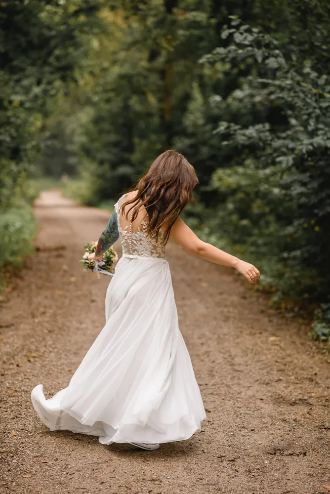 Woman in white wedding dress walks down a dirt path, holding bouquet, surrounded by trees.