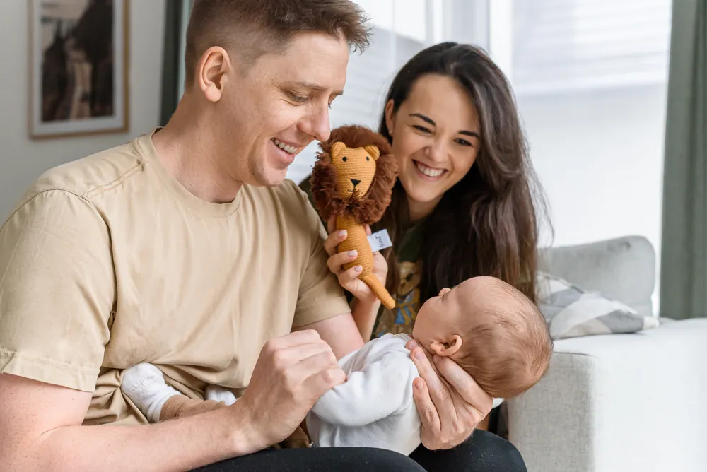 A family shares a joyful moment: a man and woman with a baby, the woman holding a lion plush toy, all smiling in a sunlit living room.