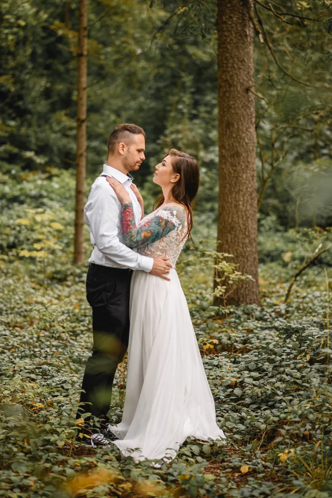Couple in forest, woman in white dress with tattoos, man in white shirt and black pants, embracing and gazing into each other's eyes.