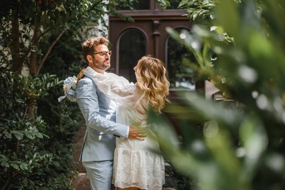 A couple in wedding attire shares an intimate moment outdoors, surrounded by lush greenery and a wooden structure, with the woman holding a bouquet.