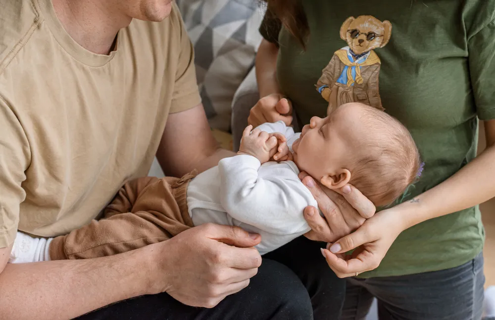 Parents tenderly cradle newborn in cozy living room; mother's bear-themed shirt and father's neutral top visible.