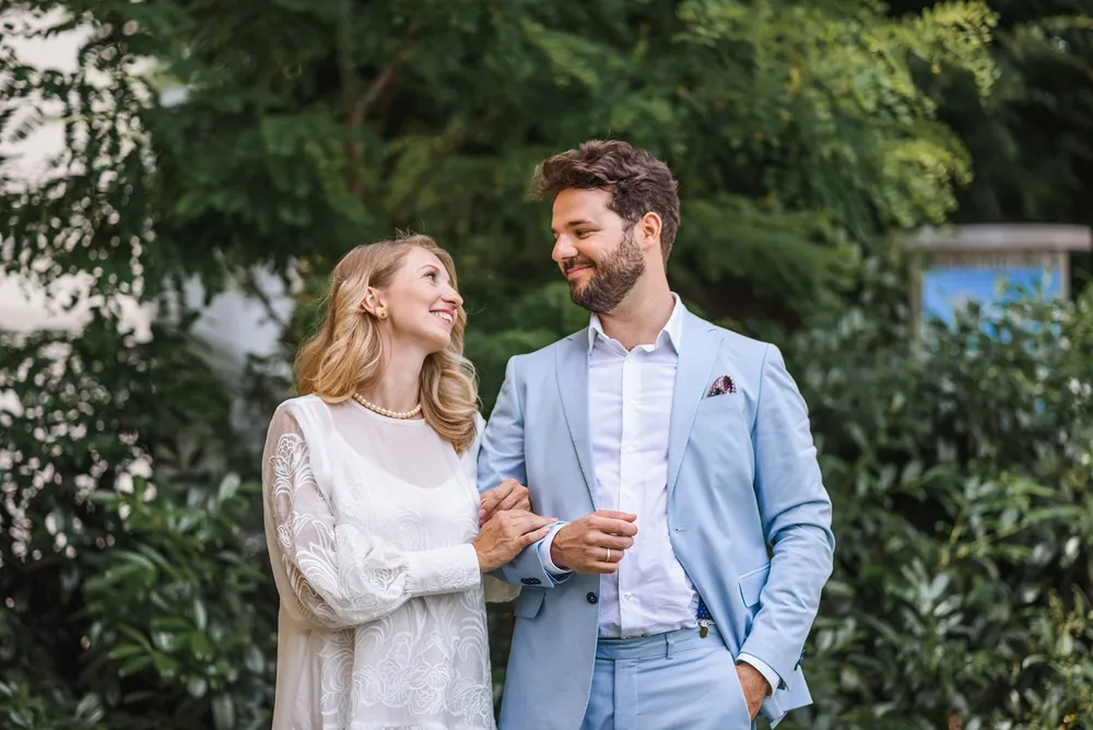 Couple in formal attire strolling in a lush garden. Woman in white lace dress, man in light blue suit. Both smiling, holding hands. Greenery and partial building facade in background.