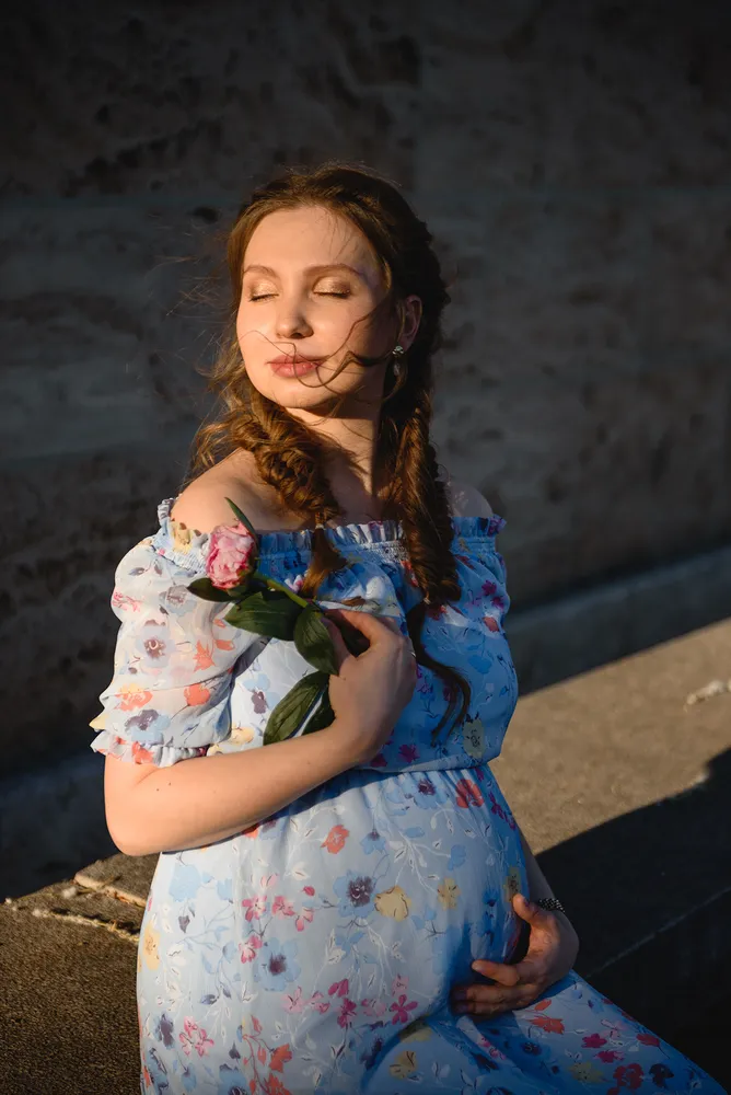 Pregnant woman in floral dress holds pink rose, eyes closed, outdoors on stone step against wall, sunlight casting shadow.