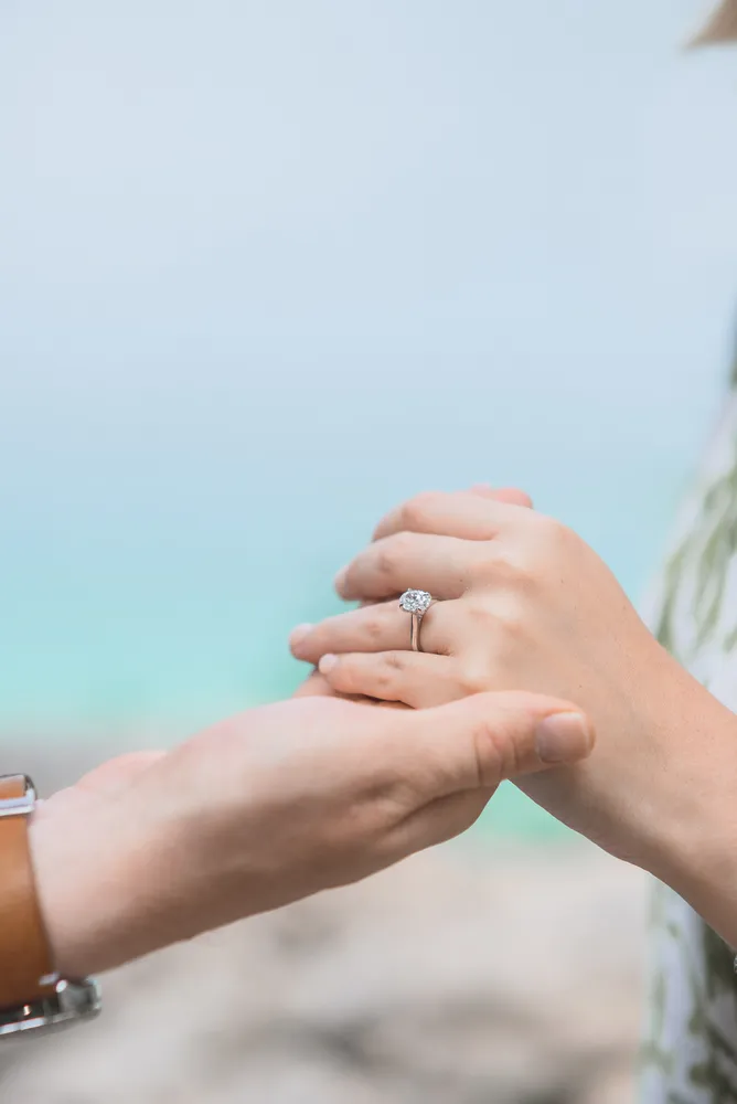 Two hands clasped together, one adorned with a diamond engagement ring, against a blurred outdoor backdrop.