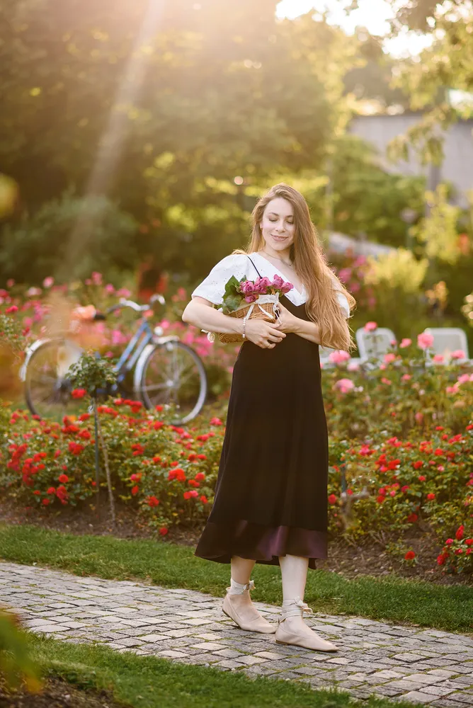 Woman in black dress holds pink flowers, walking on cobblestone path surrounded by red blooms, with a bicycle in the background.