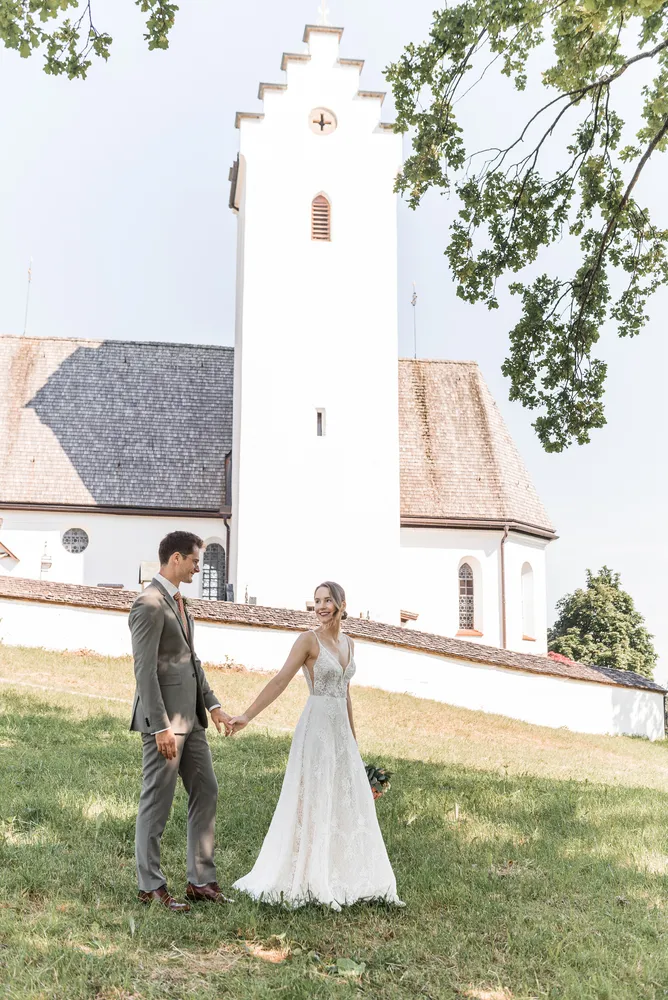 Bride in lace gown and groom in grey suit walk hand-in-hand on grassy slope toward white church with bell tower under clear sky.