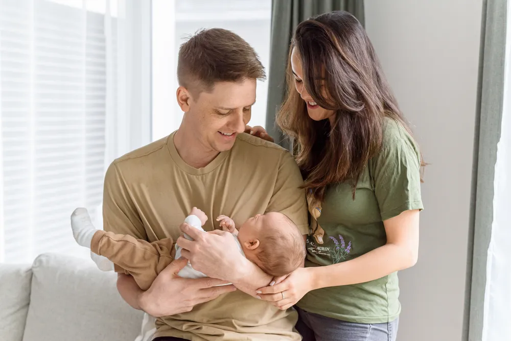 Family of three indoors; parents smiling while holding newborn on couch; soft natural light, modern decor.