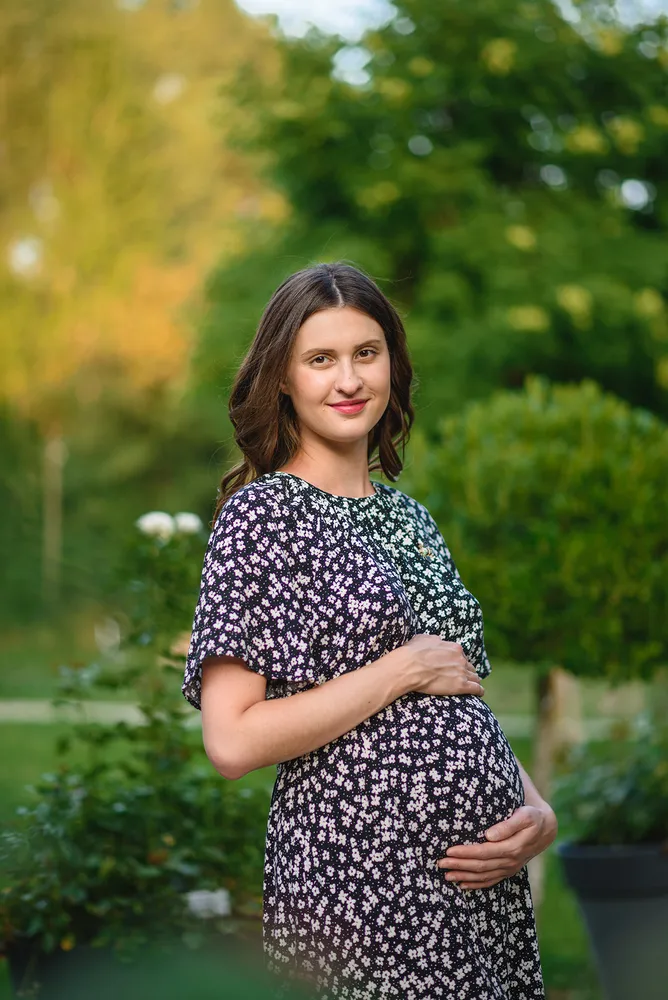 Pregnant woman in floral dress gently cradles her belly outdoors, surrounded by greenery and a hint of autumnal foliage in the background.