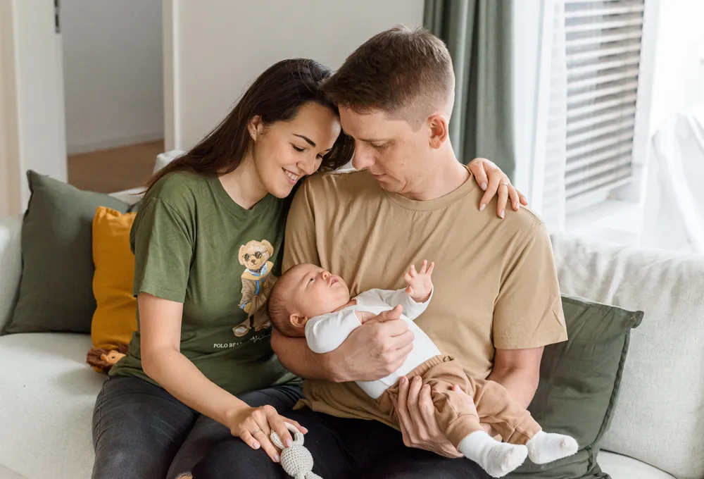 A family of three shares a tender moment on a white couch. The father cradles their baby, dressed in white and tan, while the mother smiles beside them. The baby gazes upwards, surrounded by a cozy living room with soft lighting.