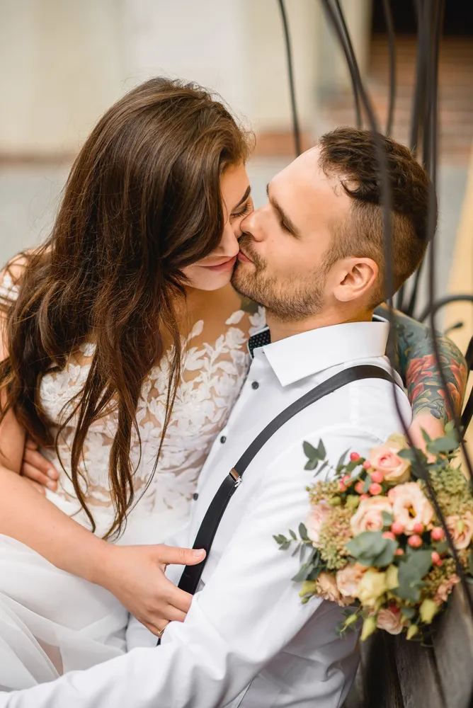 A couple shares a tender kiss; the bride in a white lace dress and the groom in a white shirt with black suspenders. He holds a bouquet of peach and white flowers. They are seated closely together, with blurred architectural details in the background, suggesting an indoor setting.