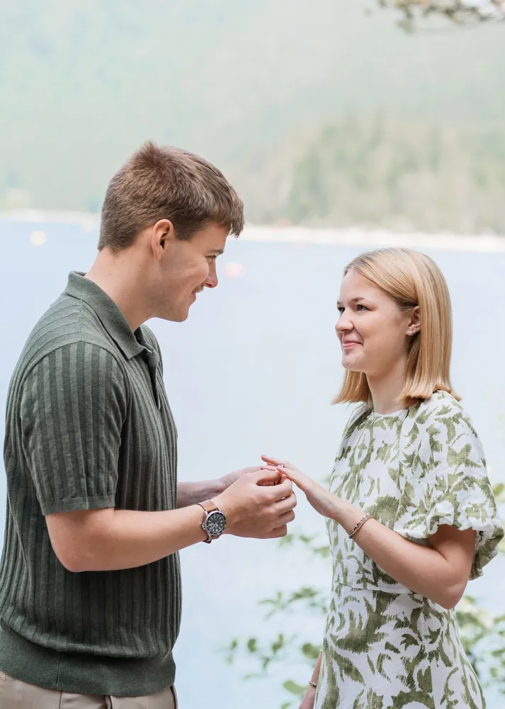 Man in olive-striped polo presents ring to smiling woman in white-green dress by serene lake, mountains in soft-focus background.
