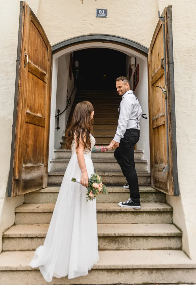 Bride descends stone steps, bouquet in hand, groom ascends beside her. They hold hands, smiling. Sunlit exterior of historic building with arched doorway and wooden doors.