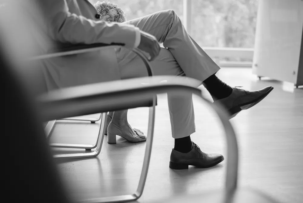 Man in suit and woman in heels sit side by side, bouquet between them, in a room with light wooden floors and a large window. Black and white photo.