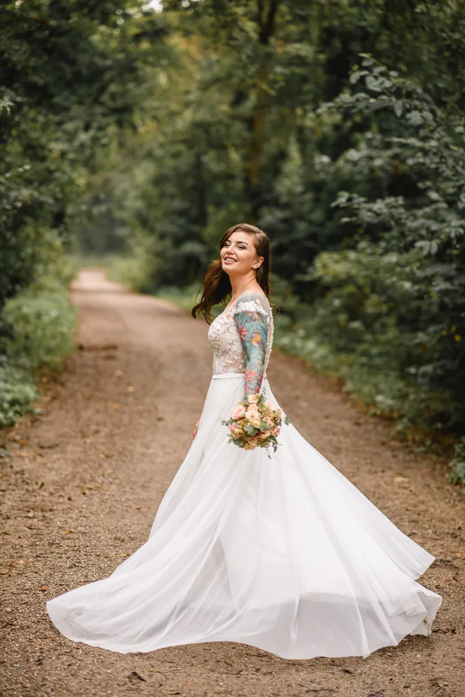 Bride in lace wedding dress with floral tattoos walks down dirt path, holding bouquet, surrounded by lush greenery.