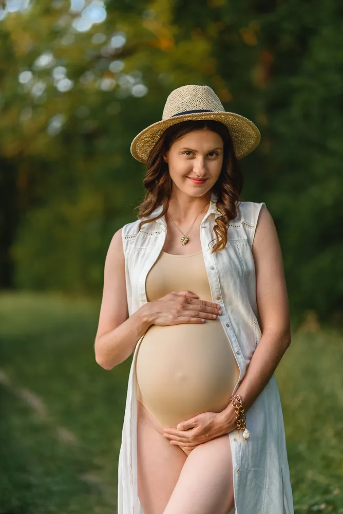 Pregnant woman in a field, wearing a straw hat and light-colored outfit, smiling while holding her belly, surrounded by greenery.