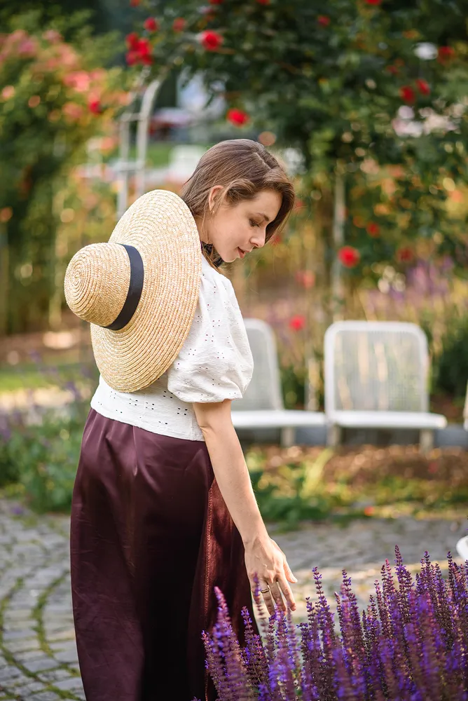 Woman in white blouse and maroon skirt, wearing a straw hat with black ribbon, tending to purple flowers in a garden setting with blurred red roses and white chairs in the background.