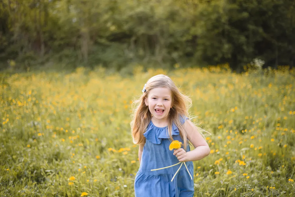 A young girl in a blue dress joyfully holds a yellow flower in a sunlit meadow, surrounded by greenery.